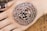 An intricately designed mehndi pattern adorns the back of a hand, featuring concentric circles with floral and spiral motifs, bordered by a detailed lace-like edge.