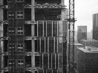 A high-rise building under construction with scaffolding, cranes, and partially completed floors. Workers can be seen on one of the upper levels of the building. In the background, there are several other tall buildings, indicating a dense urban environment.