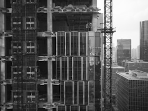 A high-rise building under construction with scaffolding, cranes, and partially completed floors. Workers can be seen on one of the upper levels of the building. In the background, there are several other tall buildings, indicating a dense urban environment.