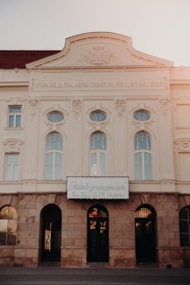 An ornately decorated building façade featuring classical architecture, with arched windows and intricate stone carvings. The upper part of the building has a decorative relief and an inscription in Latin. There is a sign in a different language below the windows.