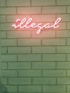 A neon sign spelling the word 'illegal' is affixed to a brick wall. The glowing letters are pink, contrasting with the muted tone of the wall, which has a slight greenish hue.