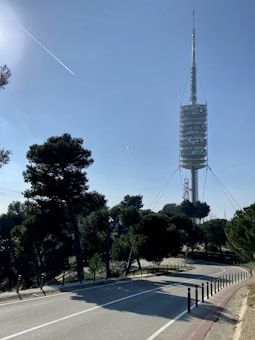 A tall, modern telecommunications tower rises into a clear blue sky. It's surrounded by green trees, and in the foreground, there's a winding road lined with posts. The overall setting appears to be a hilly area with a serene atmosphere.