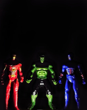 Three superhero action figures are positioned side by side against a dark background. The figure on the left is in red with gold accents, the middle figure is green with black highlights, and the figure on the right is in blue with red and white details.