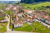 Aerial view of a historic town with a fortified church at the center, surrounded by traditional houses with red-tiled roofs. The town is set in a lush green landscape with hills in the background. People are seen gathering in an open grassy area near the church, with several cars parked along the streets.