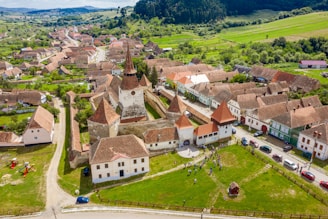 Aerial view of a historic town with a fortified church at the center, surrounded by traditional houses with red-tiled roofs. The town is set in a lush green landscape with hills in the background. People are seen gathering in an open grassy area near the church, with several cars parked along the streets.