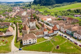 Aerial view of a historic town with a fortified church at the center, surrounded by traditional houses with red-tiled roofs. The town is set in a lush green landscape with hills in the background. People are seen gathering in an open grassy area near the church, with several cars parked along the streets.