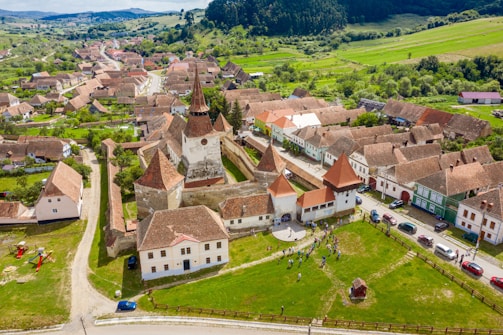 Aerial view of a historic town with a fortified church at the center, surrounded by traditional houses with red-tiled roofs. The town is set in a lush green landscape with hills in the background. People are seen gathering in an open grassy area near the church, with several cars parked along the streets.