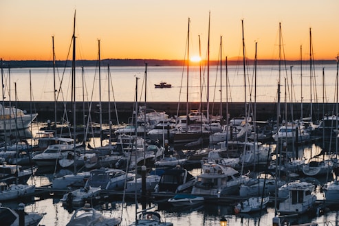 A marina filled with numerous sailboats and yachts at sunset. The sun is setting on the horizon, casting a warm orange glow across the water and sky. The masts of the boats create vertical lines against the serene backdrop.