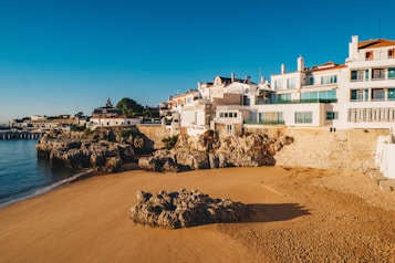 Picturesque coastal scene featuring a sandy beach bordered by rocky cliffs. Elegant white buildings with terracotta roofs and balconies overlook the beach, capturing the warmth of the sunlight. The deep blue sea meets the shore with gentle waves, adding to the tranquility of the setting. A clear blue sky enhances the serene and inviting atmosphere.