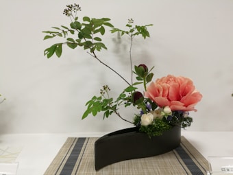 An elegant floral arrangement featuring a large, peach-colored flower prominently displayed in a black, modern-style vase. The composition includes several green leaves and small buds extending upwards, adding a sense of height and balance. A woven mat lies beneath the vase, complementing the natural and simple aesthetic of the arrangement.