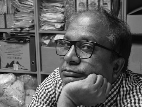 A person wearing glasses with a thoughtful expression rests their chin on their hand. Shelves filled with various stacks of papers and boxes form the background, creating a cluttered yet organized atmosphere.