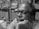 A person wearing glasses with a thoughtful expression rests their chin on their hand. Shelves filled with various stacks of papers and boxes form the background, creating a cluttered yet organized atmosphere.