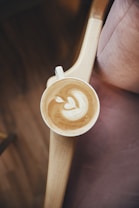 A cup of latte art with a creamy heart design rests on a wooden armrest. The surrounding setting features a warm, cozy ambiance with a soft fabric, possibly a cushion or chair, partially visible.