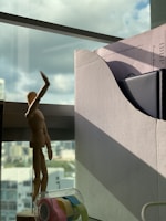 A wooden mannequin is posed with one arm raised in front of a window. Next to it is a cardboard file holder containing papers and a black folder. In the foreground, there is a transparent tape dispenser with colored tapes. The background shows an urban landscape through the window, with cloudy skies.