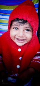A child with bright eyes and a wide smile, wearing a red knit sweater and hood with white buttons. The background features colorful horizontal stripes in blue, white, and green.