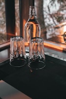 A glass bottle with a resealable cap and two upside-down drinking glasses are placed on a dark surface near a window. Warm light streams through, casting reflections and creating a cozy atmosphere.
