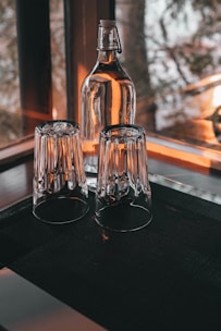 A glass bottle with a resealable cap and two upside-down drinking glasses are placed on a dark surface near a window. Warm light streams through, casting reflections and creating a cozy atmosphere.