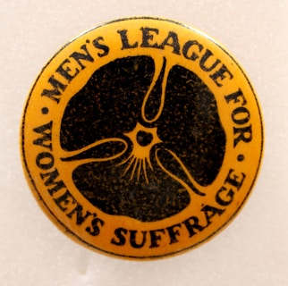 A round, vintage button with the text 'MEN'S LEAGUE FOR WOMEN'S SUFFRAGE' surrounding a central abstract design. The button has a warm, yellow-orange background with black text and design elements.