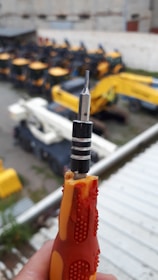 A close-up of a hand holding a screwdriver with a worn orange and yellow handle. In the background, there are multiple construction vehicles with prominent yellow and black coloring, slightly out of focus.