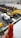 A close-up of a hand holding a screwdriver with a worn orange and yellow handle. In the background, there are multiple construction vehicles with prominent yellow and black coloring, slightly out of focus.