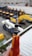 A close-up of a hand holding a screwdriver with a worn orange and yellow handle. In the background, there are multiple construction vehicles with prominent yellow and black coloring, slightly out of focus.