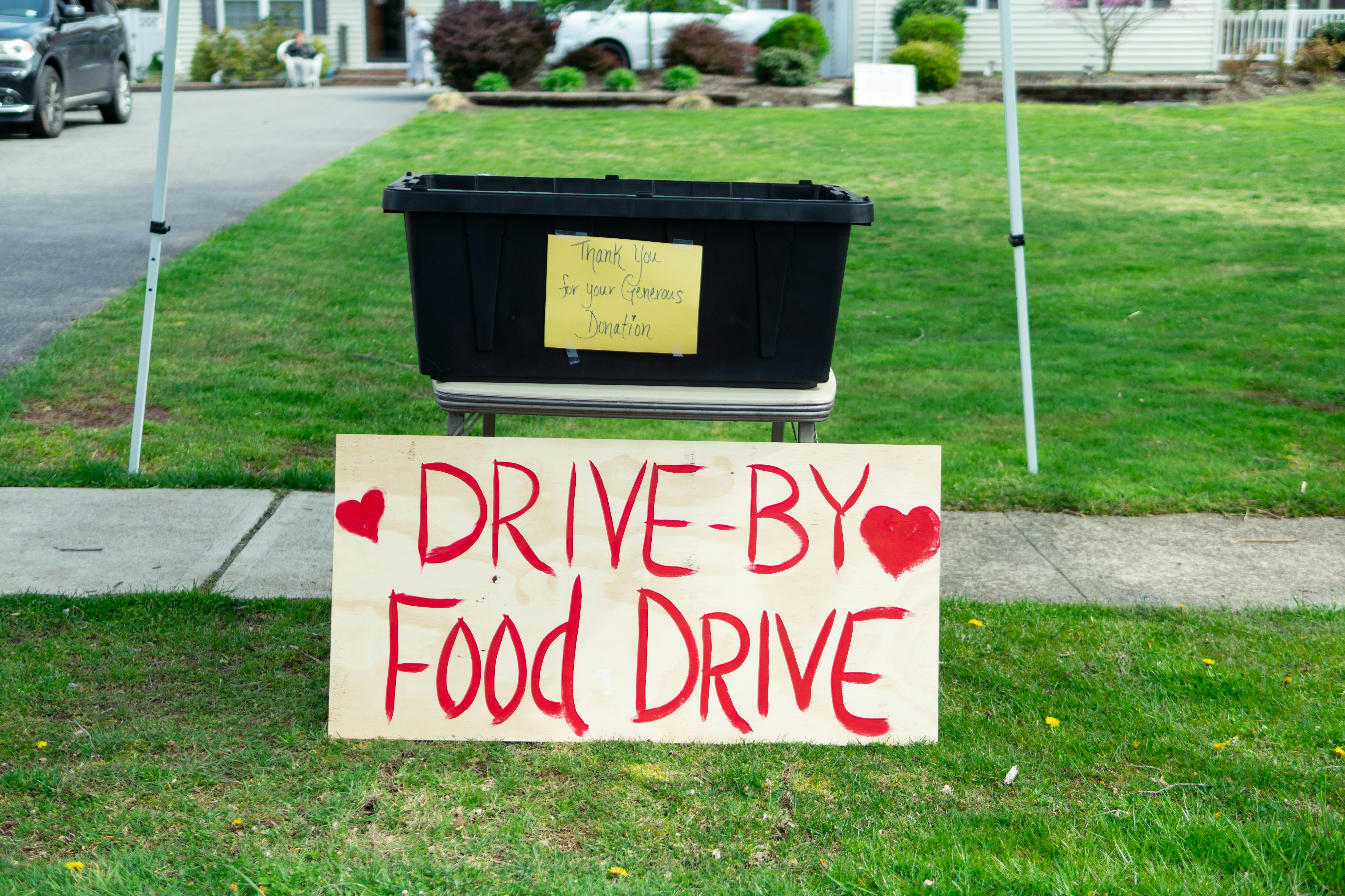 A black plastic bin is placed on a table with a yellow sign that reads 'Thank you for your generous donation.' In front of it, a large wooden sign with red lettering announces a 'Drive-By Food Drive' with hearts on either side. The setup is outdoors on a grassy area with a sidewalk and part of a residential driveway visible.