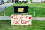 A black plastic bin is placed on a table with a yellow sign that reads 'Thank you for your generous donation.' In front of it, a large wooden sign with red lettering announces a 'Drive-By Food Drive' with hearts on either side. The setup is outdoors on a grassy area with a sidewalk and part of a residential driveway visible.