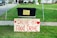 A black plastic bin is placed on a table with a yellow sign that reads 'Thank you for your generous donation.' In front of it, a large wooden sign with red lettering announces a 'Drive-By Food Drive' with hearts on either side. The setup is outdoors on a grassy area with a sidewalk and part of a residential driveway visible.