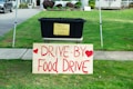 A black plastic bin is placed on a table with a yellow sign that reads 'Thank you for your generous donation.' In front of it, a large wooden sign with red lettering announces a 'Drive-By Food Drive' with hearts on either side. The setup is outdoors on a grassy area with a sidewalk and part of a residential driveway visible.