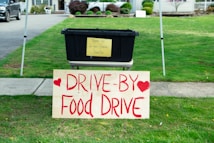 A black plastic bin is placed on a table with a yellow sign that reads 'Thank you for your generous donation.' In front of it, a large wooden sign with red lettering announces a 'Drive-By Food Drive' with hearts on either side. The setup is outdoors on a grassy area with a sidewalk and part of a residential driveway visible.