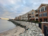 A row of modern, multi-story beachfront homes with large windows and balconies overlooking a rocky shore. The overcast sky adds a moody atmosphere, with a hint of sunset on the horizon. Waves gently lap at the pebbled beach, and a black iron fence runs alongside a paved walkway.