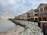 A row of modern, multi-story beachfront homes with large windows and balconies overlooking a rocky shore. The overcast sky adds a moody atmosphere, with a hint of sunset on the horizon. Waves gently lap at the pebbled beach, and a black iron fence runs alongside a paved walkway.