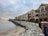 A row of modern, multi-story beachfront homes with large windows and balconies overlooking a rocky shore. The overcast sky adds a moody atmosphere, with a hint of sunset on the horizon. Waves gently lap at the pebbled beach, and a black iron fence runs alongside a paved walkway.
