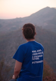 A person with short brown hair is wearing a blue t-shirt with white text printed on the back. The text includes phrases about jobs, prices, and a future, along with a hashtag. The individual is sitting on a hillside, gazing at a view of mountains and a soft, pinkish sky.