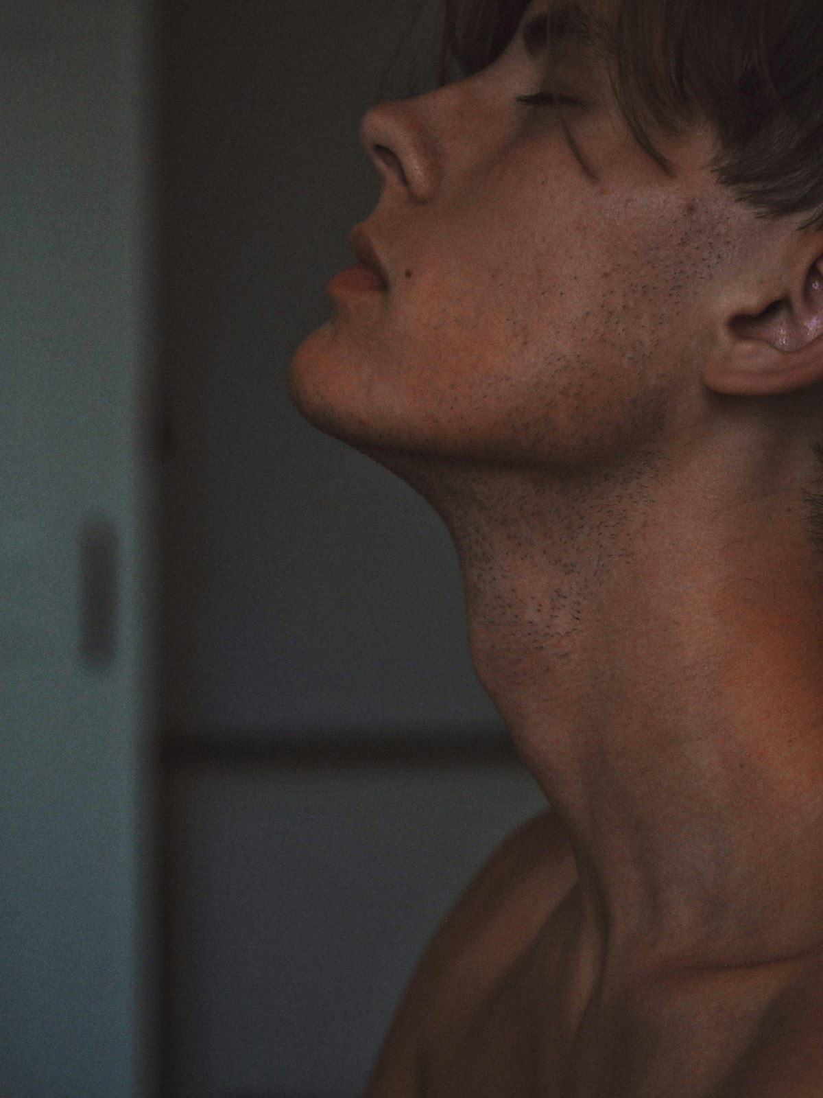 Bare torso against a pale wall, mid-breath, natural light, eyes closed.