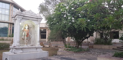 An elegant marble murti of Lord Shiva resting against a backdrop of green plants and soft sunlight.