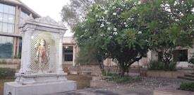 A white marble statue of a Hindu deity stands elegantly within a decorative, columned shrine. The shrine is set amidst a garden area with lush green foliage and blooming white flowers. A large modern building with glass windows is visible in the background.