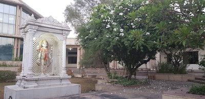 A white marble statue of a Hindu deity stands elegantly within a decorative, columned shrine. The shrine is set amidst a garden area with lush green foliage and blooming white flowers. A large modern building with glass windows is visible in the background.