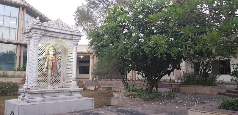 A white marble statue of a Hindu deity stands elegantly within a decorative, columned shrine. The shrine is set amidst a garden area with lush green foliage and blooming white flowers. A large modern building with glass windows is visible in the background.