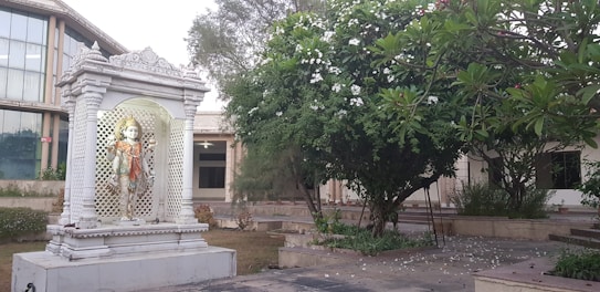 A white marble statue of a Hindu deity stands elegantly within a decorative, columned shrine. The shrine is set amidst a garden area with lush green foliage and blooming white flowers. A large modern building with glass windows is visible in the background.