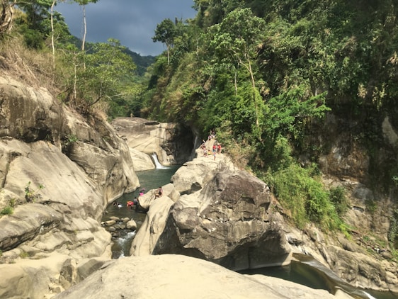 A scenic view of a rocky canyon with lush greenery surrounding it. Clear water flows through the rocks, and a group of people is seen exploring the area, some lounging on the rocks.