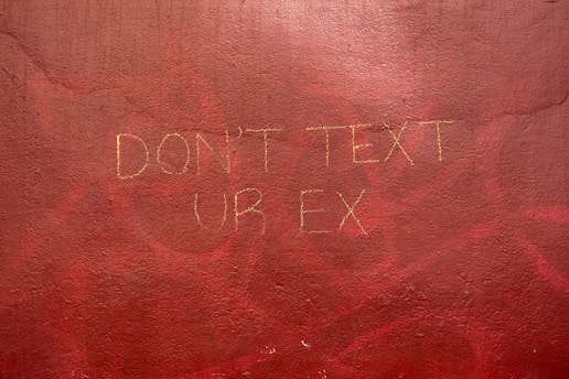 A textured wall with a hand-painted message in a faint yellow hue that reads 'DON'T TEXT UR EX'. The background is a rich, uneven red with areas of darker and lighter shades, suggesting an urban or artistic setting.