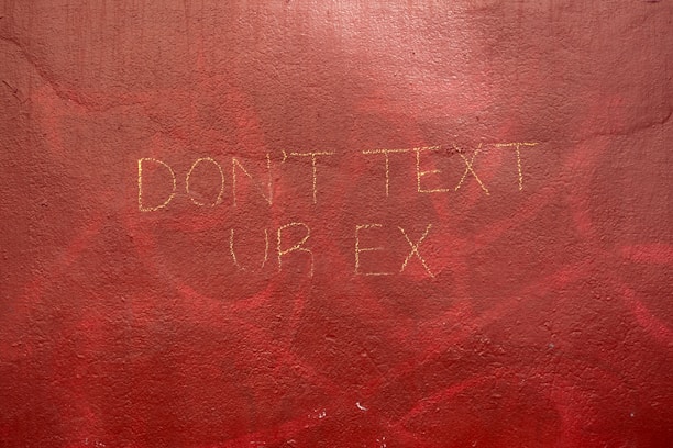 A textured wall with a hand-painted message in a faint yellow hue that reads 'DON'T TEXT UR EX'. The background is a rich, uneven red with areas of darker and lighter shades, suggesting an urban or artistic setting.