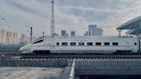 A sleek high-speed train is traveling on railway tracks with a backdrop of a modern urban skyline. The train is white with blue accents and features streamlined design, suggesting efficiency and speed. Overhead electric wires and a platform roof can be seen, indicating a railway station environment.