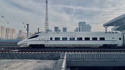 A sleek high-speed train is traveling on railway tracks with a backdrop of a modern urban skyline. The train is white with blue accents and features streamlined design, suggesting efficiency and speed. Overhead electric wires and a platform roof can be seen, indicating a railway station environment.