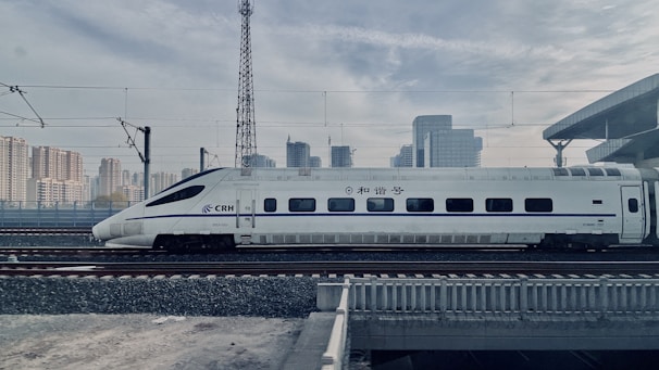 A sleek high-speed train is traveling on railway tracks with a backdrop of a modern urban skyline. The train is white with blue accents and features streamlined design, suggesting efficiency and speed. Overhead electric wires and a platform roof can be seen, indicating a railway station environment.