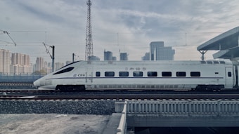 A sleek high-speed train is traveling on railway tracks with a backdrop of a modern urban skyline. The train is white with blue accents and features streamlined design, suggesting efficiency and speed. Overhead electric wires and a platform roof can be seen, indicating a railway station environment.