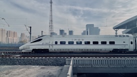 A sleek high-speed train is traveling on railway tracks with a backdrop of a modern urban skyline. The train is white with blue accents and features streamlined design, suggesting efficiency and speed. Overhead electric wires and a platform roof can be seen, indicating a railway station environment.