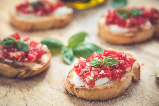 Several pieces of toasted bread with a topping of diced tomatoes and fresh basil leaves are arranged on a wooden surface. The toppings are vibrant and colorful, with bits of cheese visible beneath the tomatoes. The overall presentation is rustic and appetizing.