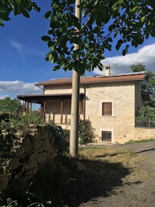 A stone house with a red-tiled roof is surrounded by greenery under a bright blue sky. A section of a wide stone wall and an unpaved path leading to the house are visible. Lush leaves from a tree dominate the upper part of the image, partially obscuring a utility pole in front of the house.